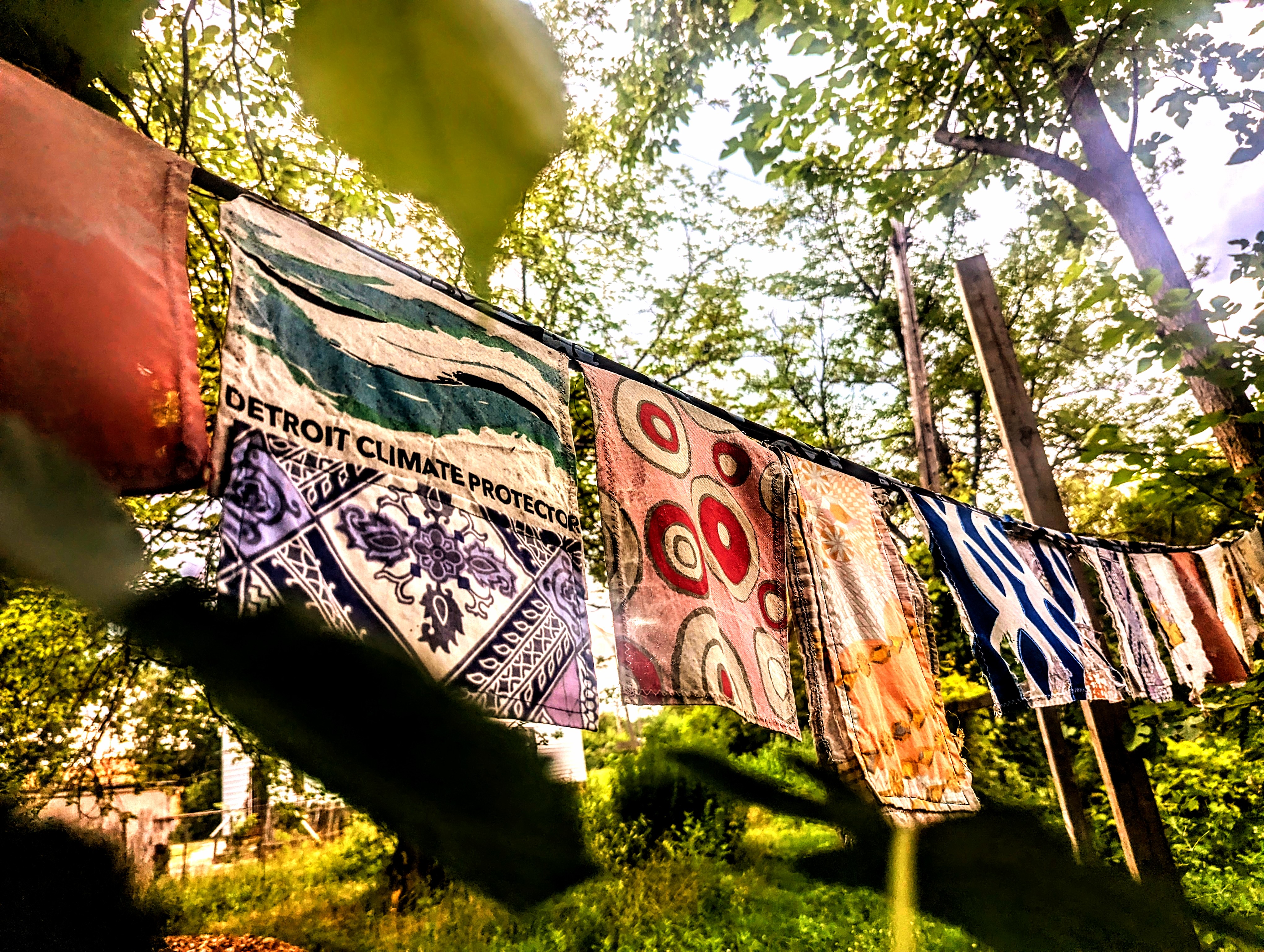 A photograph of colorful hand-dyed fabric hanging on a clothesline surrounded by trees. The camera focuses on a piece of fabric emblazoned with the phrase, "Detroit Climate Protection," and the image is overlaid with a saturated yellow hue.